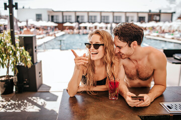Young hot couple resting at swimpool. Woman and man sit at bar and drink cocktails. Girl point to side and smile. Guy looks at her. Enjoy rest or summer vacation at swimming pool. Spa resort time.
