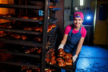 Fototapeta na wymiar Girl baker holds a tray with hot pastries in the bakery. Production of bakery products. Fresh crispy pastry rack