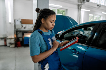 Vehicles excellence. Young african american woman, professional female mechanic using tablet pc, while standing near blue car with open hood at auto repair shop