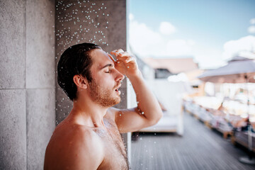 Young man taking shower outside. Side view portrait of guy without shirt standing under shower while water falling down. Enjoy water procedure.