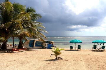 Mahahual sandy beach with green palm trees, fisher boat laying on the side and two turquoise beach umbrellas with sunbeds, sunshine, grey cloudy sky