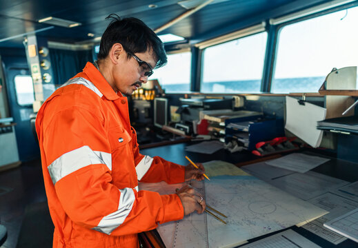 Filipino deck Officer on bridge of vessel or ship wearing coverall during navigaton watch at sea . He is plotting position on chart