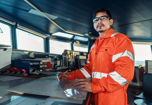 Filipino deck Officer on bridge of vessel or ship wearing coverall during navigaton watch at sea . He is plotting position on chart