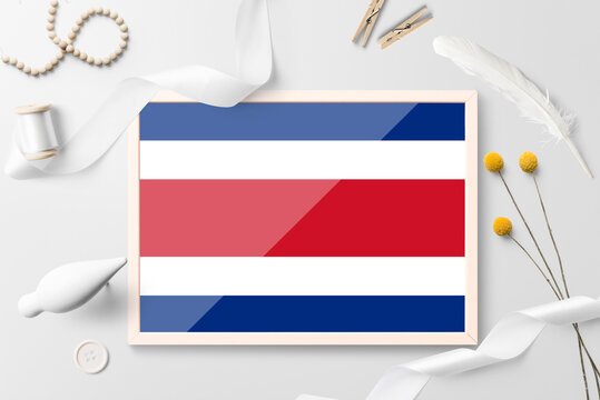 Costa Rica flag in wooden frame on white creative background. White theme, feather, daisy, button, ribbon objects.