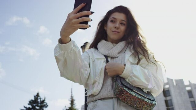 A bottom view slow motion shot with a camera floating of a dark-haired young woman in white clothes taking photos of herself on the smartphone while standing in the park
