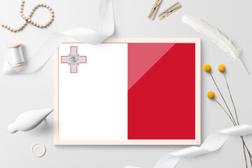 Malta flag in wooden frame on white creative background. White theme, feather, daisy, button, ribbon objects.