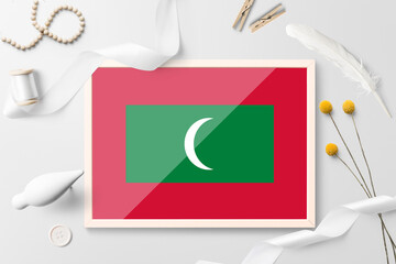 Maldives flag in wooden frame on white creative background. White theme, feather, daisy, button, ribbon objects.