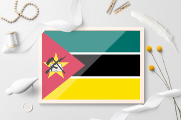 Mozambique flag in wooden frame on white creative background. White theme, feather, daisy, button, ribbon objects.
