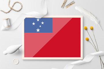 Samoa flag in wooden frame on white creative background. White theme, feather, daisy, button, ribbon objects.