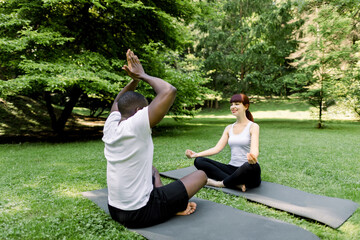 Morning yoga exercises and healthy lifestyle. Multiethnic couple doing yoga in park together, sitting in yoga pose on yoga mats outdoor at sunny summer day in park. Training and meditation