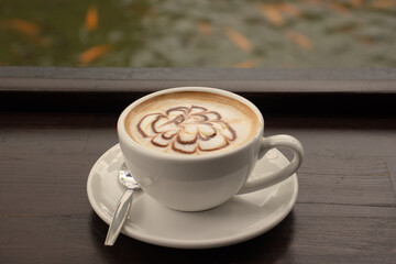 Beautiful close up of freshly brewed coffee on a wooden bench with faded goldfish in a green fish pond in the background