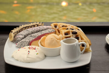Vanilla ice cream, whipped cream, fruit and waffles close up on a wooden bench with faded goldfish in a green fishpond in the background