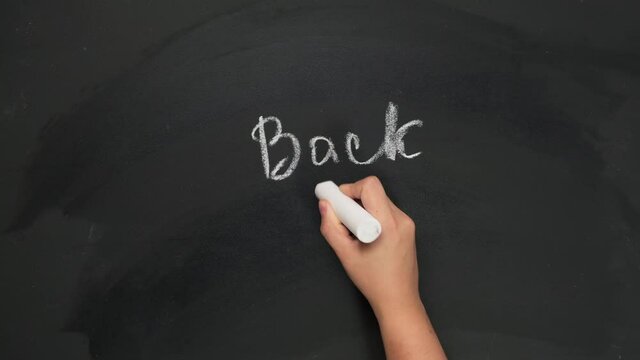 female hand holds white chalk and writes back to school on black chalk board, start of the school year