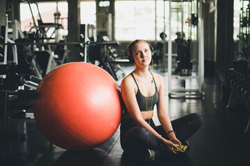 Fototapeta na wymiar A beautiful woman wearing a sports shirt is sitting and relaxing by listening to music from headphones. With rubber ball, yoga or exercise ball