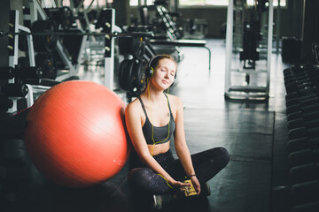 A beautiful woman wearing a sports shirt is sitting and relaxing by listening to music from headphones. With rubber ball, yoga or exercise ball