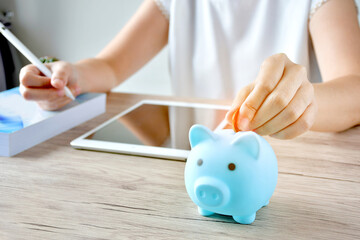 A woman puts a coin in a blue piggy bank on a wooden table to save money And record household income on the tablet.