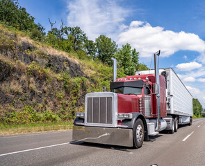 Big rig classic dark red semi truck with stylish accessories transporting load in refrigerator semi trailer driving on the straight road with rock hill in sunny day