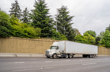 Big rig white bonnet day cab semi truck transporting commercial cargo in semi trailer moving on the highway road with concrete wall on the side