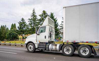 Compact powerful big rig white day cab semi truck with dry van semi trailer running on the wide highway road with trees on the side