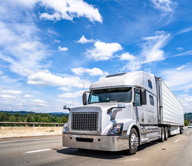 Fototapeta na wymiar Gray big rig semi truck with chrome grille transporting load in refrigerator semi trailer running on the wide highway road with clouds sky