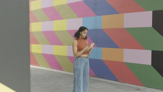 Pretty Generation Z Afro American woman influencer using smart phone on street with colourful wall 