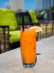 Bright orange health drink with slice of lemon set on gray table against background of modern chairs with lime green fabric and blue sky with skyscrappers in the distance. 