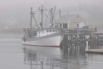Fototapeta premium Calm, restful scene in coastal Maine. Fishing trawler anchored alongside dock and enveloped in thick fog at picturesque fishing village of Port Clyde.