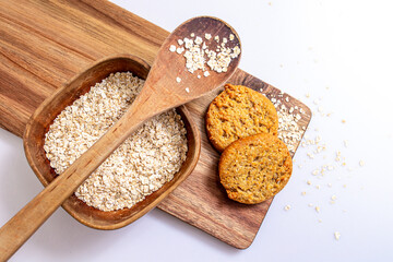 Homemade cookies with oatmeal on wooden board with a wooden container with oats flakes, and wooden spoon, over white background.