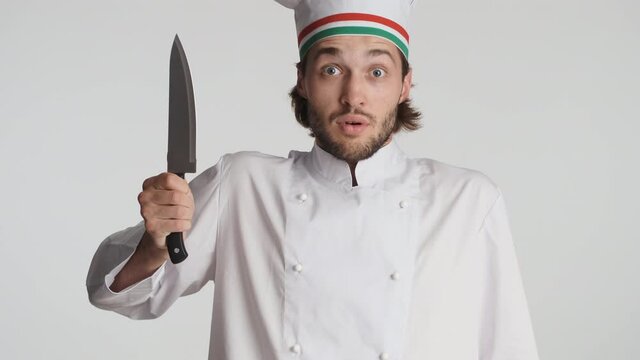Attractive italian chef in uniform holding knife amazedly looking in camera over white background. Wow expression