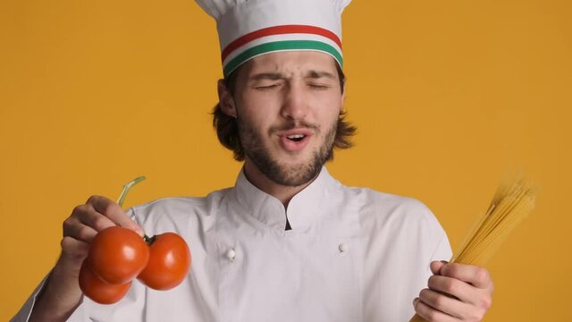 Portrait of handsome italian chef in uniform holding fresh tomatoes and macaroni happily singing over colorful background