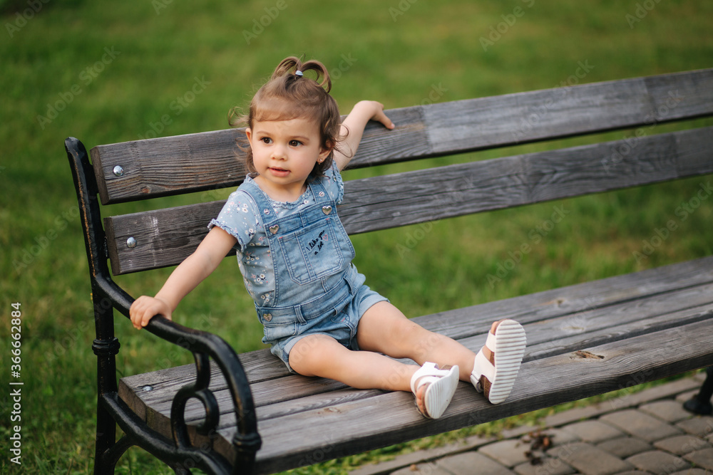 Wall mural cute little girl in denim climbs on the bench in the park. happy smiled kid on the bench