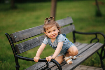 Cute little girl in denim climbs on the bench in the park. Happy smiled kid on the bench