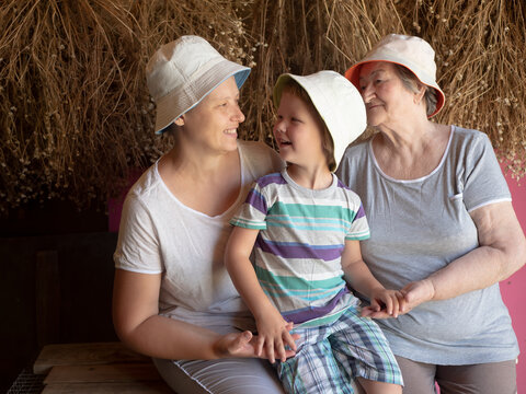 Beautiful Elderly Woman With Wrinkles, Young Girl And Boy From Same Family. Three Different Ages Of Similar People. Mother, Adult Daughter And Grandson In Identical Hats On Background Of Dry Flowers