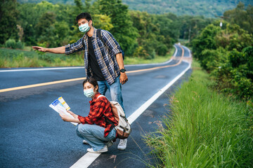 Female tourists sit and look at the map, male tourists pretending to hitchhike. Both wear masks and...