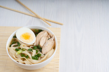 Spicy asian ramen soup made of chicken broth with noodles decorated by halved egg, meat slices and green onion in a bowl standing on a bamboo mat on white wooden table. Image with copy space