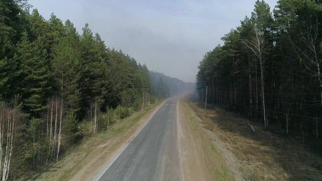 Aerial view of a forest road and smoke from a forest fire. Cars drive along the road. There is a fire engine near the fire. Summer, hot sunny day.
