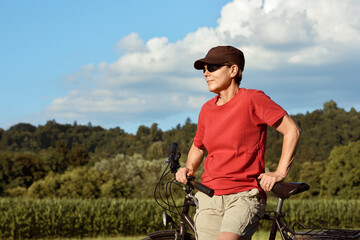 A portrait of a middle-aged female cyclist in a red summer T-shirt resting next to her bike.