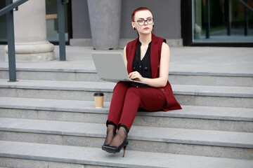 Fototapeta na wymiar Young girl in a red jacket sitting on the steps and working at a computer. The concept of freelance work