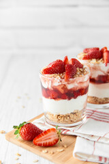 Homemade layered dessert with fresh strawberries, cream cheese or yogurt, granola and strawberry jam in glasses on white wood background. Healthy organic breakfast or snack concept. Selective focus.