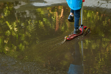 The child rides a scooter in puddles. Baby in blue T-shirt and dark pants moves puddles