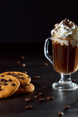 Glass cup of coffee with whipped cream and chocolate on it, chocolate chip cookies and roasted coffee beans on dark background. Concept of ready to eat food, tasty snack. Selective focus, copy space.