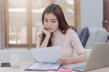 The concept of working from home, a beautiful Asian woman is working with a face that shows stress and anxiety. Looking at the white paper in the hand on the desk is a new normal form of work.