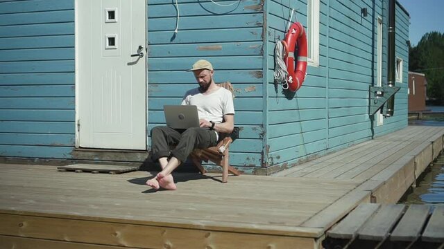 Man Opening Laptop And Start Working Spbd. Male On Lake Works Remote With Wireless Computer. Bearded Hipster Guy Sit By Water Lake. Vacation Of Freelancer. Houseboat Hotel Cabin