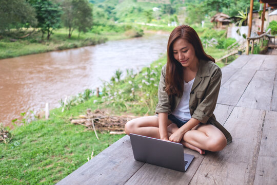 Portrait image of a beautiful asian woman working on laptop computer while sitting by the river with mountains and nature background