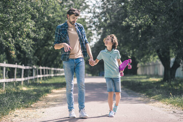 Dark-haired boy with a skateboard and his father walking in the park