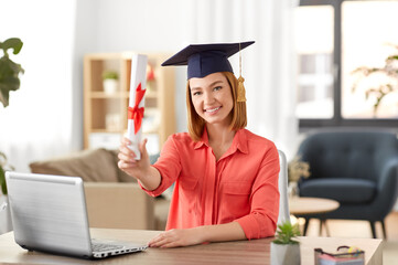 e-learning, education and people concept - happy smiling female graduate student in mortarboard with laptop computer and diploma at home