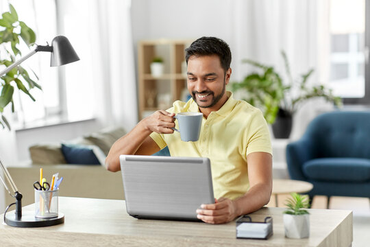 technology, remote job and lifestyle concept - happy indian man with laptop computer drinking coffee or tea at home office