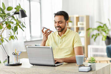 technology, communication and people concept - happy smiling indian with smartphone, earphones and laptop computer using voice command recorder at home office