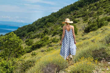 Fototapeta na wymiar Young woman wearing striped summer dress and straw hat walking among super bloom of wildflowers, relaxing while enjoing beautiful nature of of Adriatic sea coastal nature of Croatia.
