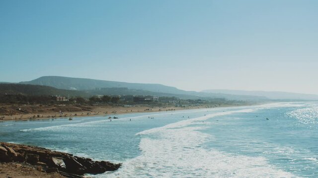 Surfing in Atlantic Ocean, Morocco, many people on the beach, big waves and clear sky and mountains on horizon, Taghazout village, 4k